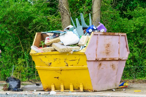 A large yellow and pink plastic skip positioned on a gravel surface, filled with assorted household waste including plastic storage containers, fabric items, cardboard boxes, and miscellaneous rubbish, with some items leaning over the edge of the skip. The background features a backdrop of green foliage and trees, indicating an outdoor environment. The skip is placed next to a paved area, and a small black trash bag is visible on the ground nearby. The scene suggests a waste collection or clearance operation, typical of rubbish removal services such as those offered by Business Waste Removal Pinner, serving local areas around the postcode regions near Pinner in London.