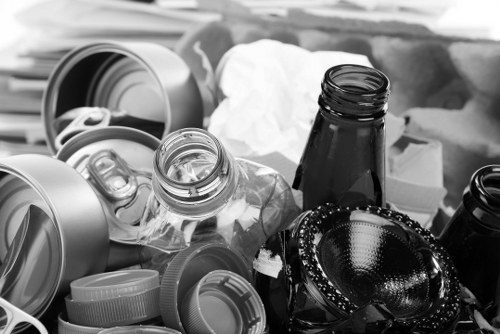 A collection of mixed waste includes several aluminium cans, some with open tops and others flattened, along with glass bottles, one of which is a dark-coloured beer or soda bottle, all placed disorderly on a surface. There are plastic bottle caps scattered among the debris, and a few crumpled pieces of paper or tissue are visible in the background. The bottles and cans are in varying conditions, with some appearing clean and others with minor dents or scratches. The scene suggests an indoor environment, possibly a countertop or table, with a focus on the waste items awaiting collection or disposal. This image aligns with the services offered by Business Waste Removal Pinner, highlighting the typical types of rubbish handled during waste clearance activities in the local area, such as Pinner or surrounding parts of northwest London, ensuring relevance to local rubbish removal needs.