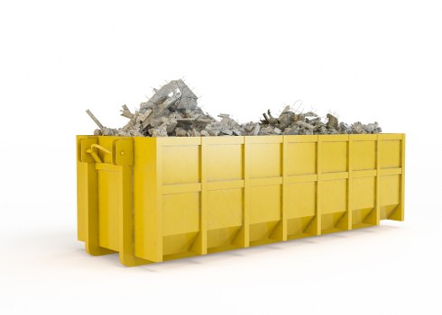 A large, yellow industrial skip positioned outdoors on a paved surface, filled with a mixture of broken concrete, brick debris, and construction rubble. The skip features a solid metal construction with a ribbed exterior and a sturdy, enclosed design to contain loose materials. It is situated against a plain, white background, emphasizing the full load of mixed waste materials spilling over the top edge. The debris includes irregularly shaped concrete chunks, some with rough, textured surfaces, and smaller fragments of brick and mortar, indicating construction or renovation waste typical of a site in Pinner or the surrounding northwest London area. The skip's side panel has visible hinges and reinforcement bars, with the shape of the load slightly rising in the middle due to the uneven distribution of debris. This image visually represents a common waste disposal solution provided by companies such as Business Waste Removal Pinner, specializing in rubbish collection and disposal services for commercial clients.