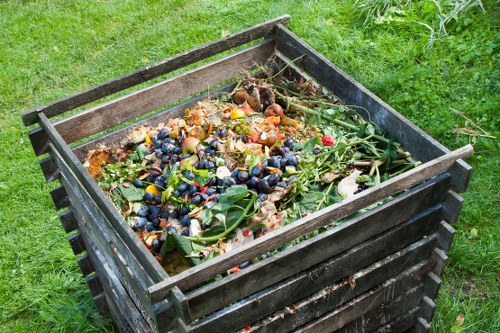 A black wooden compost bin filled with mixed garden waste, including fruit and vegetable scraps, green leaves, and flower trimmings, situated on a green grassy outdoor area. The bin is rectangular with slatted sides, positioned on a paved space adjacent to a lush, well-maintained lawn, with some grass visible at the base of the bin. The waste inside appears partially decomposed and is overflowing slightly at the top. The environment suggests a backyard or garden setting suitable for waste collection and removal services, and the image conveys the types of garden refuse managed by a business waste removal provider in Pinner. The overall scene emphasizes the importance of proper waste disposal in a residential garden context, consistent with services offered by Business Waste Removal Pinner.