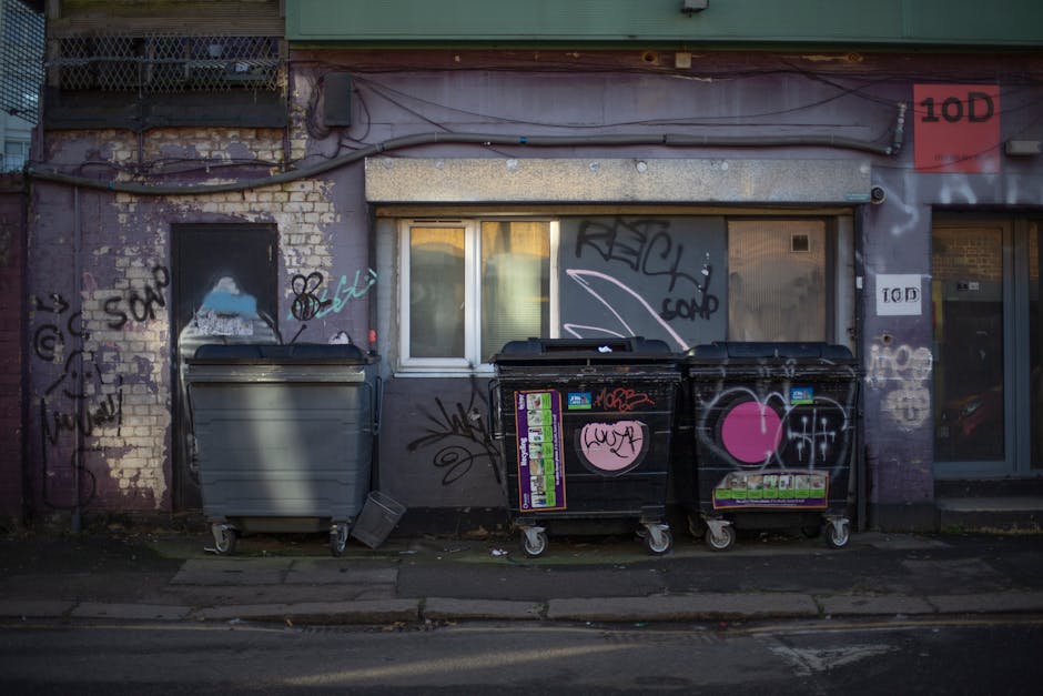 Row of four large, rectangular orange waste containers positioned against a tall, smooth blue metal wall. The containers have a rugged, weathered appearance with visible dirt, rust, and black graffiti